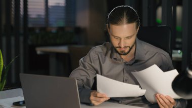 Akşamları bürodaki Kafkasyalı işadamı evrakları kontrol ediyor veri bilgilerini rapor ediyor dizüstü bilgisayar evraklarını daktilo ediyor iş adamının memnun olduğu şirket muhasebecisi şirket maliyesini kontrol ediyor.