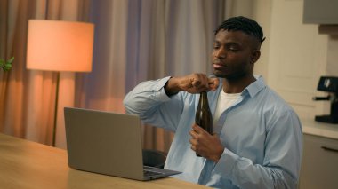 African American man at kitchen male relaxing video call online conference guy alcohol addiction drinking beer bottle home evening bad habit laptop looking at computer watching sport game alcoholism