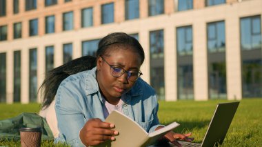Afro-Amerikan mutlu kadın üniversite akademisi genç kız lisesi parkta yeşil çimlere uzanmış kitap telif defteri bilgisayarlarıyla çalışıyor sınav sınavlarına hazırlanmak için kahve içiyor