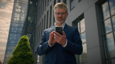 Gülümseyen orta yaşlı işadamı şehrin dışında ofis yakınında cep telefonu kullanıyor kameraya gülümse mutlu yetişkin iş adamı işvereni akıllı telefon simsarı emlakçısı