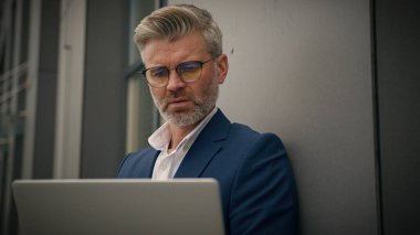 Kafkasyalı yaşlı, olgun, işletme yöneticisi, orta yaşlı, işveren, duvar binasına dayanan, dizüstü bilgisayarda çalışan, yorgun, çalışmaktan yorulan, dışarı fırlayan camlar.