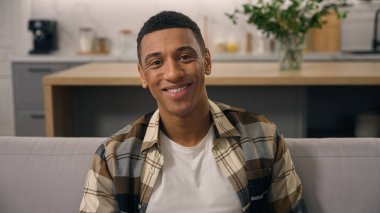 Evde rahatlayan etnik adamın portresi. Kapalı alanda gülümseyen bir adam. Kendi evindeki rahat kanepeye bakıyor.