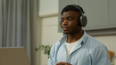 African American man user gamer guy male at home kitchen evening put on wireless headphones earphones with laptop computer listen music audio song prepare to work video conference game online sound