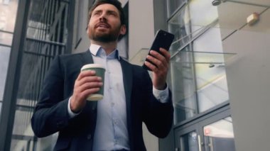 Mutlu, mutlu, orta yaşlı, beyaz, erkek, iş adamı, girişimci, elinde kahve fincanı, cep telefonu ve akıllı telefon konuşmalarıyla belediye binasına giriyor.