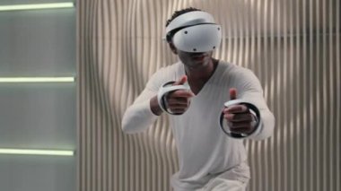 Afro-Amerikalı kaygısız erkek, 3 boyutlu sanal gerçeklik kaskı takan VR gözlük takıyor, simülasyon oynuyorlar modern metaevren oyunu oynuyorlar gelecekteki beyaz stüdyoda silah kontrolü yapıyormuş gibi davranıyorlar.