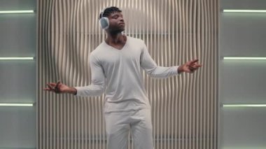 Aktif dans müziği Afrikalı Amerikalı kaygısız adam erkek neşeli dans kablosuz kulaklıklar takıyor fütüristik beyaz stüdyoda ses kalitesinde dans ritmi dinliyor.