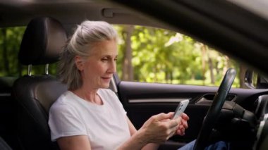 Neşeli Kafkasyalı olgun kadın yaşlı, yaşlı, yaşlı, emekli kadın, emekli araba sürücüsü, cep telefonu kullanıyor, cep telefonu kullanıyor, yumruğunu sıkıyor, zaferi kutluyor.