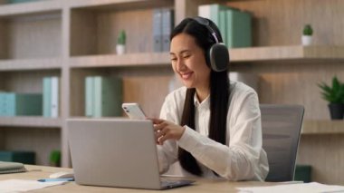 Mutlu gülümseyen Asyalı Japon iş kadını Koreli Çinli iş kadını ofis çalışanı cep telefonu akıllı telefon uygulaması kullanarak kablosuz müzik kulaklığı dinliyorlar.