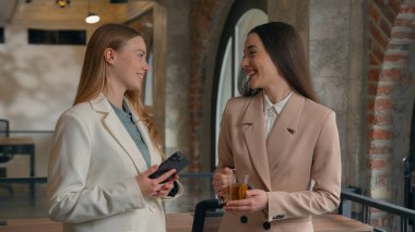 İki mutlu gülümseyen Kafkasyalı kadın ofis çalışanları iş kadınları iş kadınları iş arkadaşları iş konuşmalar sohbet konuşmalar haberler çay fincanları cep telefonu kahkahaları arkadaşlık