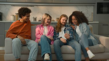 Kafkasyalı adam elinde cep telefonu tutarak çok ırklı insanlara komik bir görüntü gösteriyor. Çeşitli arkadaşlar gülüyor, neşe saçıyor, erkek Afrikalı kadınlar kanepede oturup gülüyor.