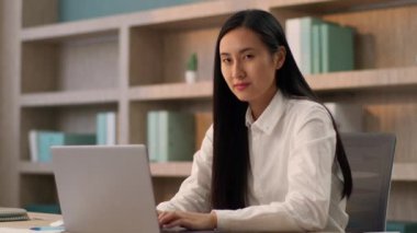 Gülümseyen güler yüzlü Japon ofis çalışanı Çinli kadın Koreli öğretmen iş kadını kız sekreter dizüstü bilgisayarda çalışıyor masa üstü bilgisayarına bakan kamera görüntülü gülücük içeride.