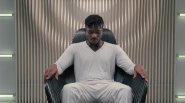 Beyaz oda fütürizm stüdyosunda gülümseyen Afrikalı etnik mutlu siber adam kameraya bakan erkek adam kaygısızca gülümsüyor bilgisayar teknolojisinde modern gelecekçi uzay teknolojisinde oturuyor.