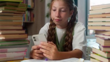 Sıkılmış, sakin, Kafkasyalı, küçük kız, liseli, öğrenci öğrenci, iki kitap yığınının arasında oturuyor. Okul kütüphanesinde cep telefonu uygulaması kullanıyor.