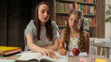 Kafkas kadın öğretmen eğitmeni, liseli küçük bir kızı işaret ederek, ilkokul öğrencisinin kitap okuduğunu, ders kitabı okuduğunu, ödev takımına yardım ettiğini ve gülümseyen özel ders öğrencisine işaret ettiğini açıklıyor.