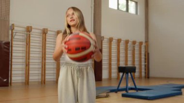 Beyazların küçük kızı basketbol topu tutuyor, spor salonu spor salonu gülümsüyor, beden eğitimi okuyor, kız öğrenci öğrenci eğitimi alıyor, spor salonu öğrencisi.