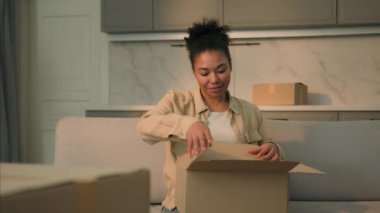 Afro-Amerikalı kadın mutlu ev sahibi kutuları açıyor karton kutuları açıyor yeni eve taşınıyor kiralanmış daireye taşınıyor kadın müşteri kız paketleri açıyor internetten alışveriş malzemeleri alıyor.
