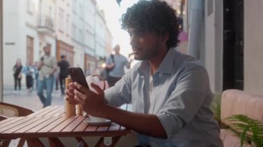 Arap latin erkek ziyaretçi kafe müşterisi mutlu müşteri işadamı açık havada cep telefonu kullanarak kahve içiyor Hintli erkek turist başparmağı gösteren seyahat uygulaması akıllı telefonu kullanıyor.