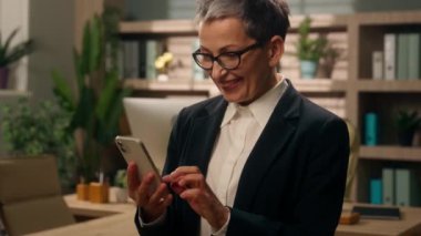 Ofiste mutlu bir şekilde gülümseyen yaşlı beyaz kadın iş kadını olgun kıdemli işveren orta yaşlı CEO girişimci cep telefonu sosyal medya akıllı telefonu kullanıyordu.