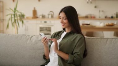 Mutlu gülümseyen, rahat Hintli kadın Arap kadın etnik kız sosyal medya akıllı telefonuna cep telefonu uygulaması teknolojisi kullanarak bakıyor evden alışveriş yaparken bağımlı zımbırtısıyla sohbet ediyor.