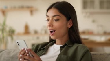 Mutlu gülümseyen pozitif şaşkın Hintli kadın Arap etnik kız sosyal medya akıllı telefonuna internette göz gezdirirken cep telefonu sohbetini evde kaydırarak iyi haber teklifini kabul et.