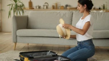 Hintli kadın Arap kadın etnik kız evde Hintli bayan yolculuk için hazırlanıyor bavul valiziyle yolculuk için şapka ve güneş gözlüğü takıp gülümseyerek eğlenceli moda tarzını şımartıyor.