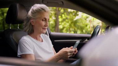 Kendinden emin, yaşlı, beyaz, kadın, kadın, emeklilik şoförü, otomobil taşıma aracında cep telefonu, akıllı telefon, cep telefonu uygulaması ve kameralı sohbet uygulaması kullanıyor.