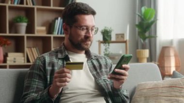 Gülümseyen Kafkasyalı yetişkin bağımlı erkek ev kanepesi ödemesinde cep telefonu alışverişi yapıyor internetten internet satın alıyor elinde kredi kartı, internet kartı, nakitsiz e-ticaret, akıllı telefon uygulaması tutuyor.