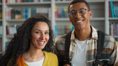 İki mutlu öğrenci Kafkas kadın Afrikalı Amerikalı erkek öğrenci kütüphanede pozitif eğitim üniversiteden arkadaşlar sınıf arkadaşları gülümsüyor lisede gülerek kameraya bakıyor.