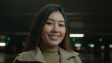 Asyalı mutlu, etnik, mutlu, mutlu, gülümseyen bir Çinli kadın güzel, Koreli, muhteşem, Japon, iş kadını, iş kadını, yeraltı otoparkında, kameranın dişlek gülüşüne bakıyor.