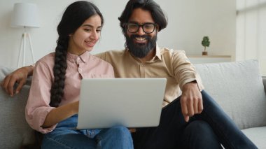 Kanepede oturan Hintli çift bilgisayar alışverişi yapıyor. İnternetten internet alışverişi yapıyor. Mutlu, çok ırklı eş, gülümseyen bir koca.