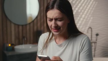 Stresli, üzgün, Kafkasyalı bir kadın. Keyifsiz, bayan kullanıcı, akıllı telefon bağımlısı, ev tuvaletinde cep telefonu ekranı görüntüsü, kötü haber mesajı, cep telefonu, internet kumar sorunu.