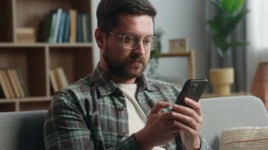 Neşeli, sakin, ciddi, rahat erkek oyuncu kullanıcısı cep telefonu akıllı telefon cihaz bağımlısı sosyal medya alışverişi online uygulama sohbet ev koltuğu bakınarak oturma odasında
