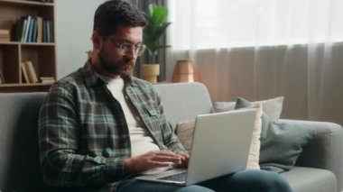 Yetişkin serbest çalışan iş adamı ev erkeği erkek yazar kullanıcı dizüstü bilgisayardaki yazı yazma makalesini kullanıyor ev kanepede çalışıyor düşünüp internet işi alışverişi yapıyor.
