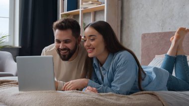Mutlu Kafkasyalı kadın ve erkek yatakta kaygısız bir şekilde yatıyorlar. Yatak odasında dizüstü bilgisayardan komik videolar izliyorlar. Gülümsüyorlar.