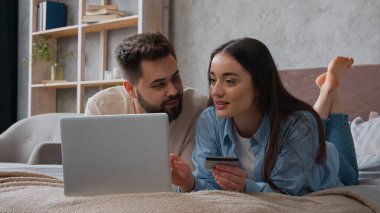 Kafkasyalı çift yatakta yan yana yatıyorlar kredi kartı kullanıyorlar dizüstü bilgisayar bankacılığı uygulamasına şifreyi giriyorlar gülümseyen kadın ve erkek alışveriş siparişleri ev yatak odasında ödeniyor.