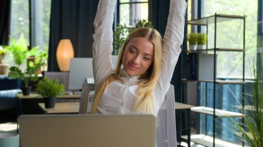 Kafkasyalı bir kadın dizüstü bilgisayarla iş projesi yazıyor bilgisayarlara bakıyor masa başında rahatlamış iş kadını çay içiyor kahve içiyor, ellerini dinlendirip sandalyeye yaslanıyor.