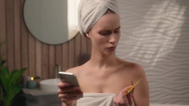 Banyo havlusuna sarılı beyaz bir kadın, elinde kredi kartı bankacılığı, ödeme sorunu, banka hesabı, tuvalet alışverişinde cep telefonlu telekız hatasını kullanarak para kaybetmeye karşı kızgın.