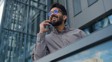 Ofis terasında cep telefonuyla konuşan neşeli Hintli bir adam mutlu gülümseyen Arap iş adamı CEO yönetici akıllı telefon şehir dostu iş konuşmasında dışarıda cep telefonu görüşmesi