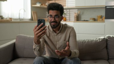 Mutlu etnik Hintli Arap erkek blogcu iş adamı, mutfaktaki rahat koltukta cep telefonuyla sohbet ediyor.