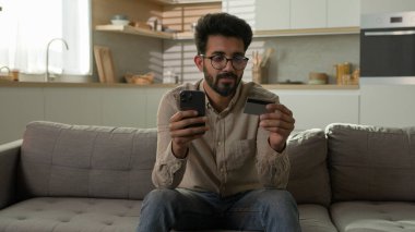 Gülümseyen Arap Etnik Erkek Alışverişçi Müşteri mutfak kanepede online alışveriş mobil mağaza uygulaması ile telefon ödeme banka kredi kartı mal satın alma sipariş para transferi e-ticaret