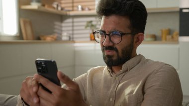 Kafası karışmış Hintli Arap bir adam. Kanepedeki erkek ev sahibi. Akıllı telefon aleti kullanıyor. Sosyal medyayı okuyor.