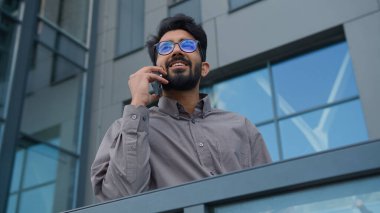 Ofis terasında cep telefonuyla konuşan neşeli Hintli bir adam mutlu gülümseyen Arap iş adamı CEO yönetici akıllı telefon şehir dostu iş konuşmasında dışarıda cep telefonu görüşmesi