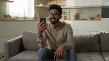 Mutlu etnik Hintli Arap erkek blogcu iş adamı, mutfaktaki rahat koltukta cep telefonuyla sohbet ediyor.