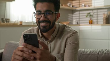 Hint Arap milenyumu çocuğu mutlu gülümseyen adam hafta sonu kanepede dinleniyor cep telefonuyla internet üzerinden kayıyor akıllı telefon uygulaması kullanıyor ev mutfağında internetten sipariş veriyor.