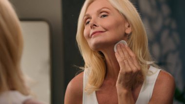 50 'li yaşlarda, güzel, olgun bir kadın yüzüne pamuk yastıkla toz ve lüks kozmetik uyguluyor. Kusursuz pürüzsüz cilt makyajı yapıyor. Aynalı ev banyosunda, yüz kırışıklıkları, yaşlanmayı önleyen güzellikler.