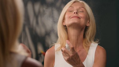 Sağlıklı gülümseyen, 50 'li yaşlardaki beyaz kadın banyo aynasında taze nemlendirici su sıkarak yaşlanma karşıtı lüks kozmetik vücut sisi spreyi makyaj spreyini tazeleyen fıskiyeli güzellik ürününü düzeltiyor.