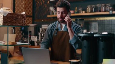 Kafkasyalı mutlu bir çalışan cep telefonuyla konuşan erkek barista müşterisiyle konuşuyor. Küçük işletme sahibi, neşeli garson, kafeterya restoranında birden fazla iş yapıyor.