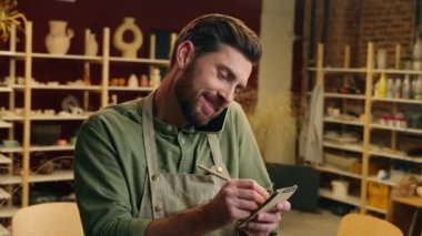 Gülümseyen Kafkasyalı erkek girişimci mutlu küçük işletme sahibi sanatçı iş adamı cep telefonlu müşteri siparişi çömlek seramik atölyesine kalem yazma emri