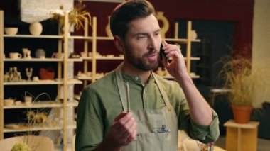 Beyaz adam, erkek girişimci, iş adamı, çömlekçi, apronlu, cep telefonlu küçük işletme sahibi, cep telefonundan konuşan, malzeme siparişi veren seramik çömlek atölyesi atölyesinde düzeni tartışan.