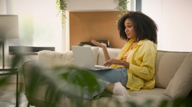 Etnik genç kız Afro-Amerikalı kadın kanepesinde dizüstü bilgisayarla video konferansında sohbet ederek neşeli 20 'li yaşlarda kadın bilgisayar başında sanal toplantı hakkında konuşuyor.